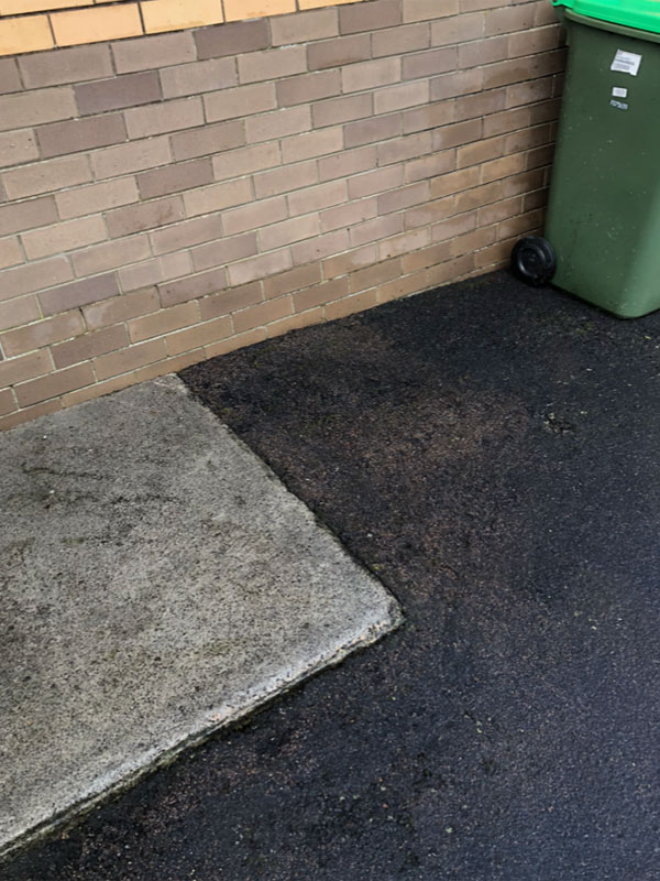 Concrete slab and dark asphalt beside a brick wall and green wheelie bin